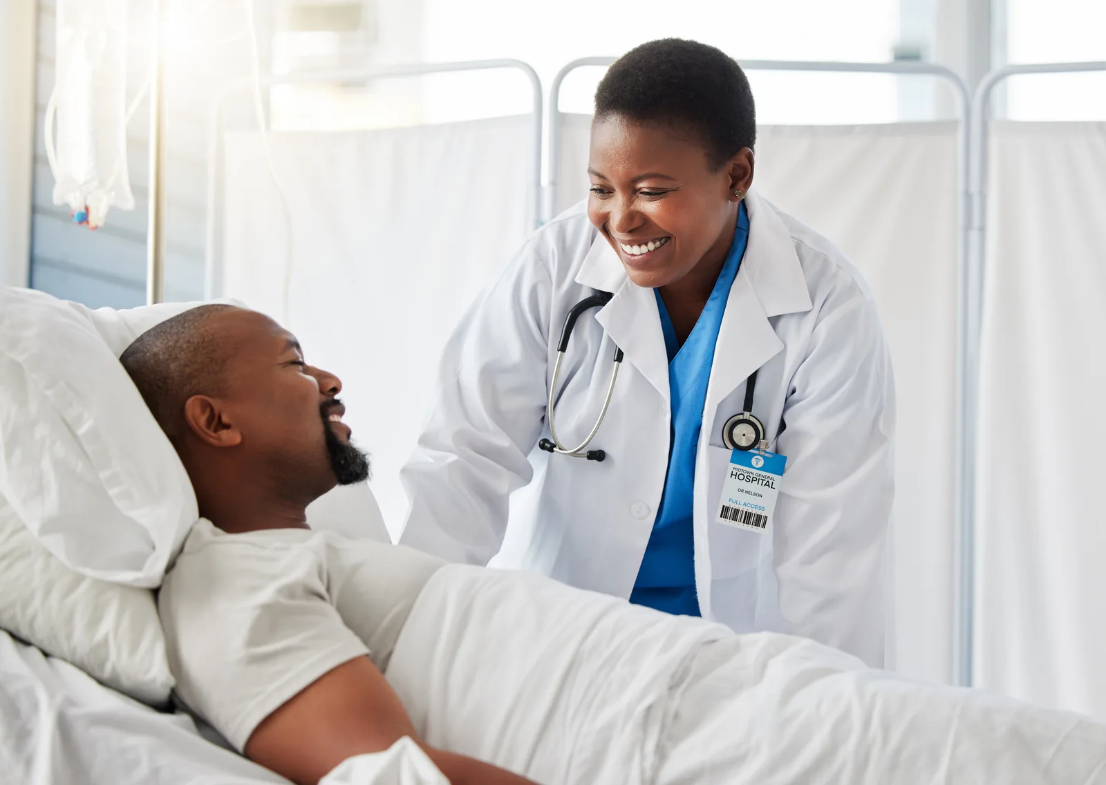 A smiling Black doctor speaking with a patient beside his hospital bed